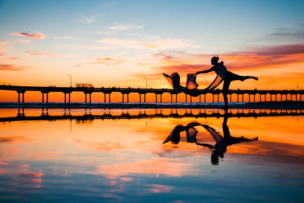 gallery-03 Silhouette of a dancer performing near a jetty at sunset with vibrant sky reflection.